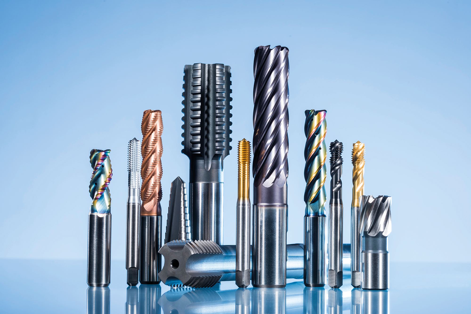 A variety of metal milling and cutting tools, including end mills and taps, are arranged upright on a reflective surface against a blue gradient background.